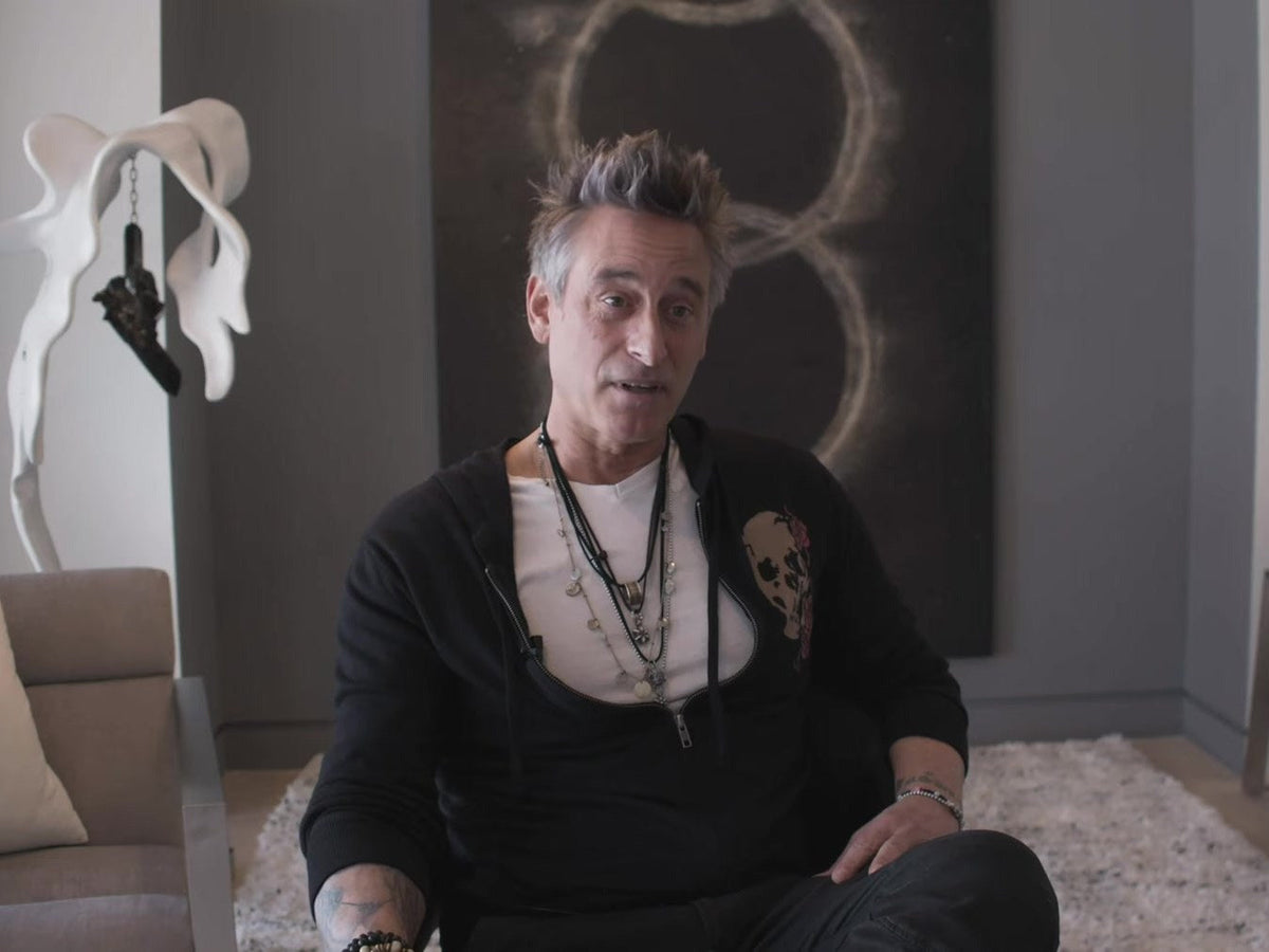 Artist Matthew Samson seated and speaking in an interview, with a large abstract painting on the wall behind him